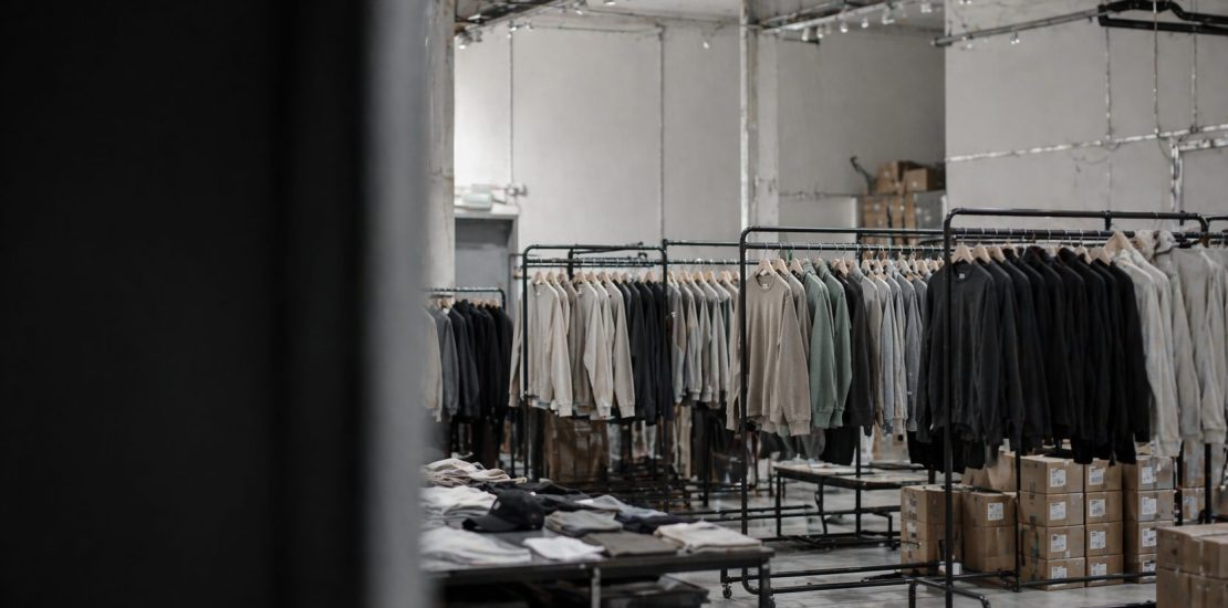 Industrial clothing storage: rows of jackets on black metal racks in a warehouse, with stacked boxes on the floor and tables of folded garments nearby.