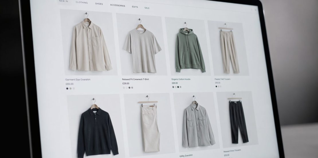 Laptop screen showing an online clothing store with a grid of six product photos (shirts, hoodie, pants) in neutral tones