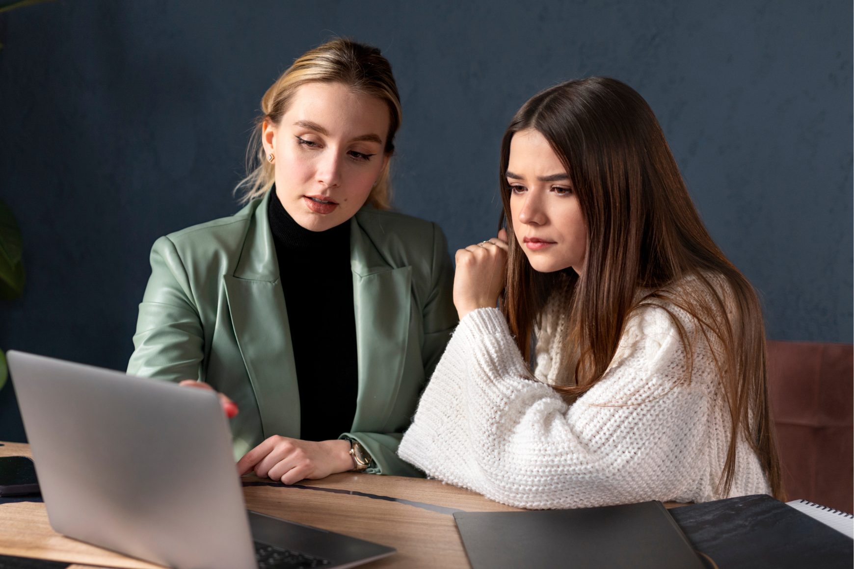 Two women sit at a table, focused on a silver laptop screen; one wears a green blazer, the other a white knit sweater, consulting together.